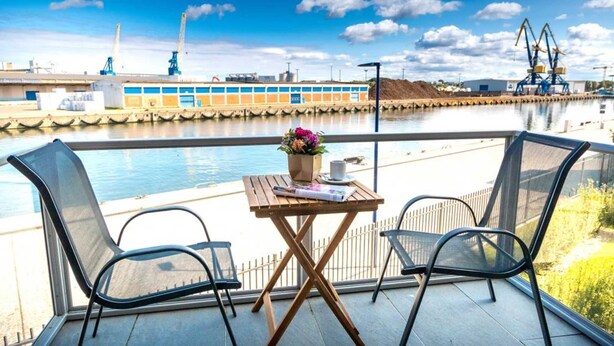 Ein Balkon mit Tisch und zwei Stühlen, mit Blick auf ein Gewässer und Industrieanlagen im Hintergrund.