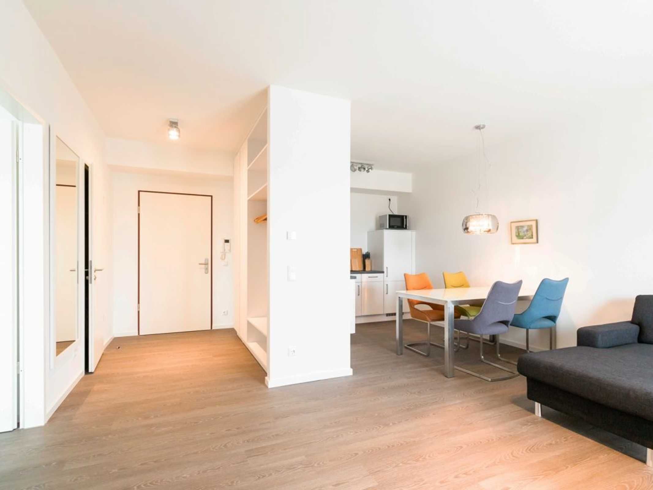 A modern apartment with a wooden floor, a dining area with a table and chairs, a sofa, and a kitchen in the background.