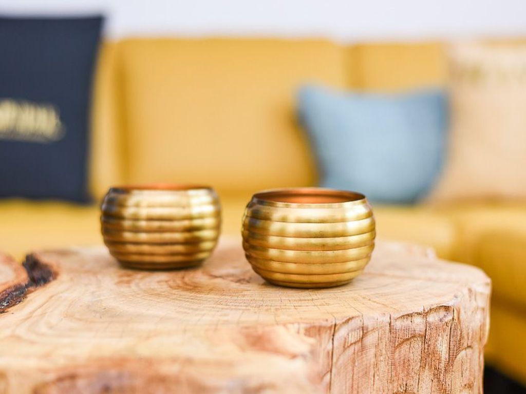 Deux vases dorés sont posés sur une surface en bois, avec un canapé jaune flou en arrière-plan.