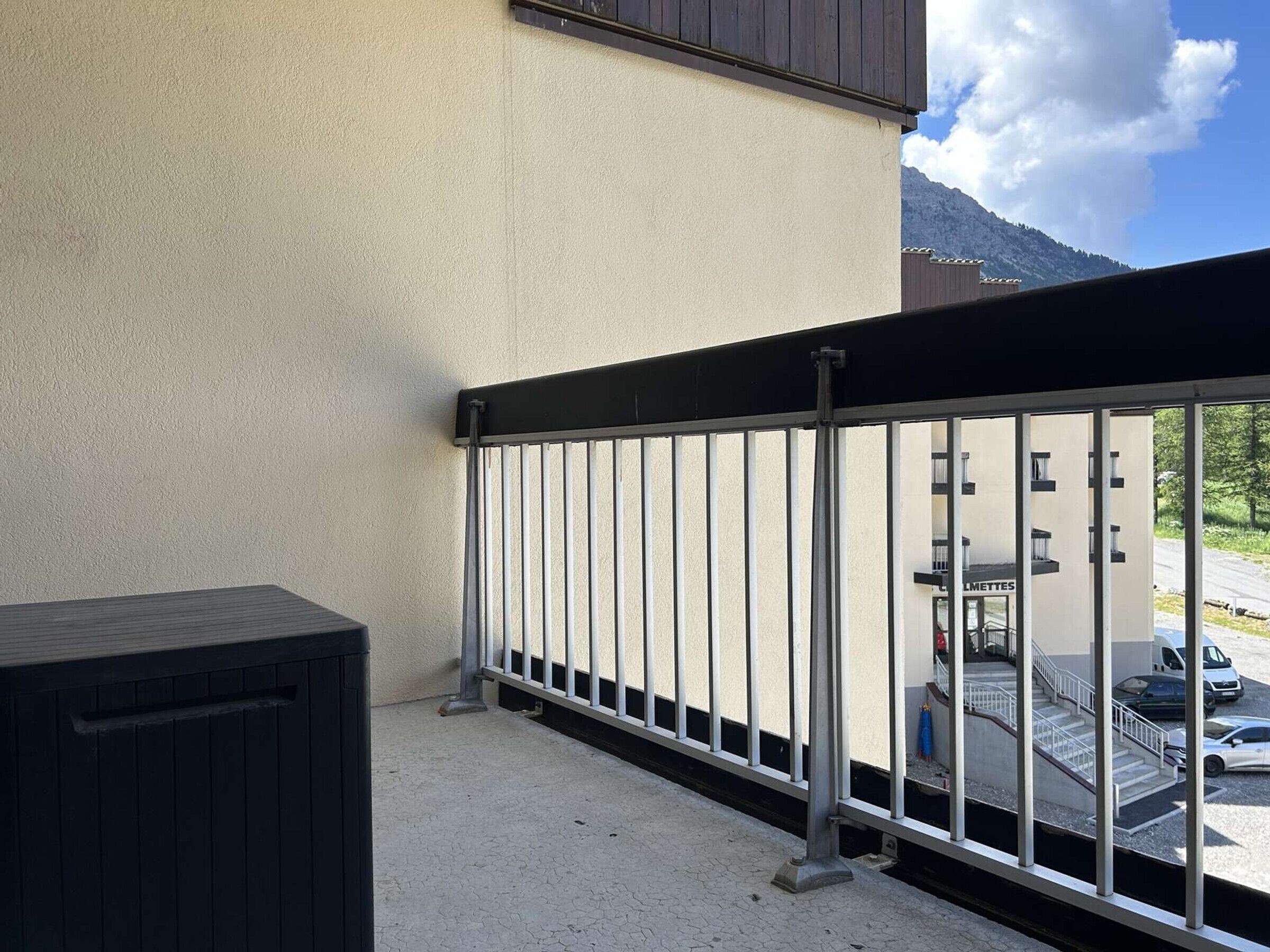 Un balcon avec une balustrade en métal, une vue sur un immeuble portant l’inscription « LIMETTES » de l’autre côté de la rue, et une montagne en arrière-plan.