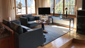 A modern living room with a sofa, TV, and fireplace. Large windows offer a view of a forested area.