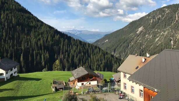 A mountain village with traditional wooden houses, a green meadow, and a forested valley.