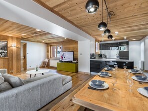Une cuisine moderne avec un plafond en bois, un coin repas avec une table en bois dressée pour quatre personnes, et un salon avec un canapé gris et un téléviseur.