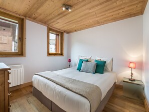 A bedroom with a wooden ceiling, a large bed with multiple pillows, a bedside table with a lamp, and a window with a view of buildings.