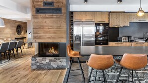 A modern kitchen with a stone fireplace, wooden cabinets, stainless steel appliances, and a bar area with stools. - Prime Ski-In/Ski-Out Location, Modern 5-Room Apartment with Fireplace (Val-d'Isère)