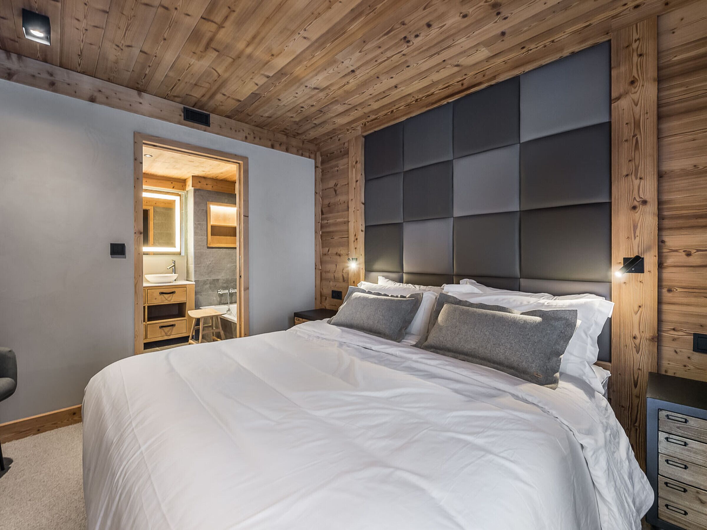 Une chambre avec un plafond en bois, un grand lit avec une literie blanche, une table de chevet et une tête de lit intégrée.