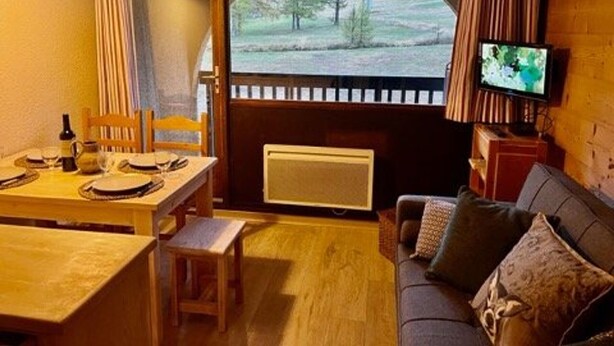 A cozy room with a dining table set for two, a sofa with cushions, a television, and a window with a view of trees and a body of water. - Apartment Montgenèvre 4 people, parking, ski-in/ski-out (Montgenèvre)