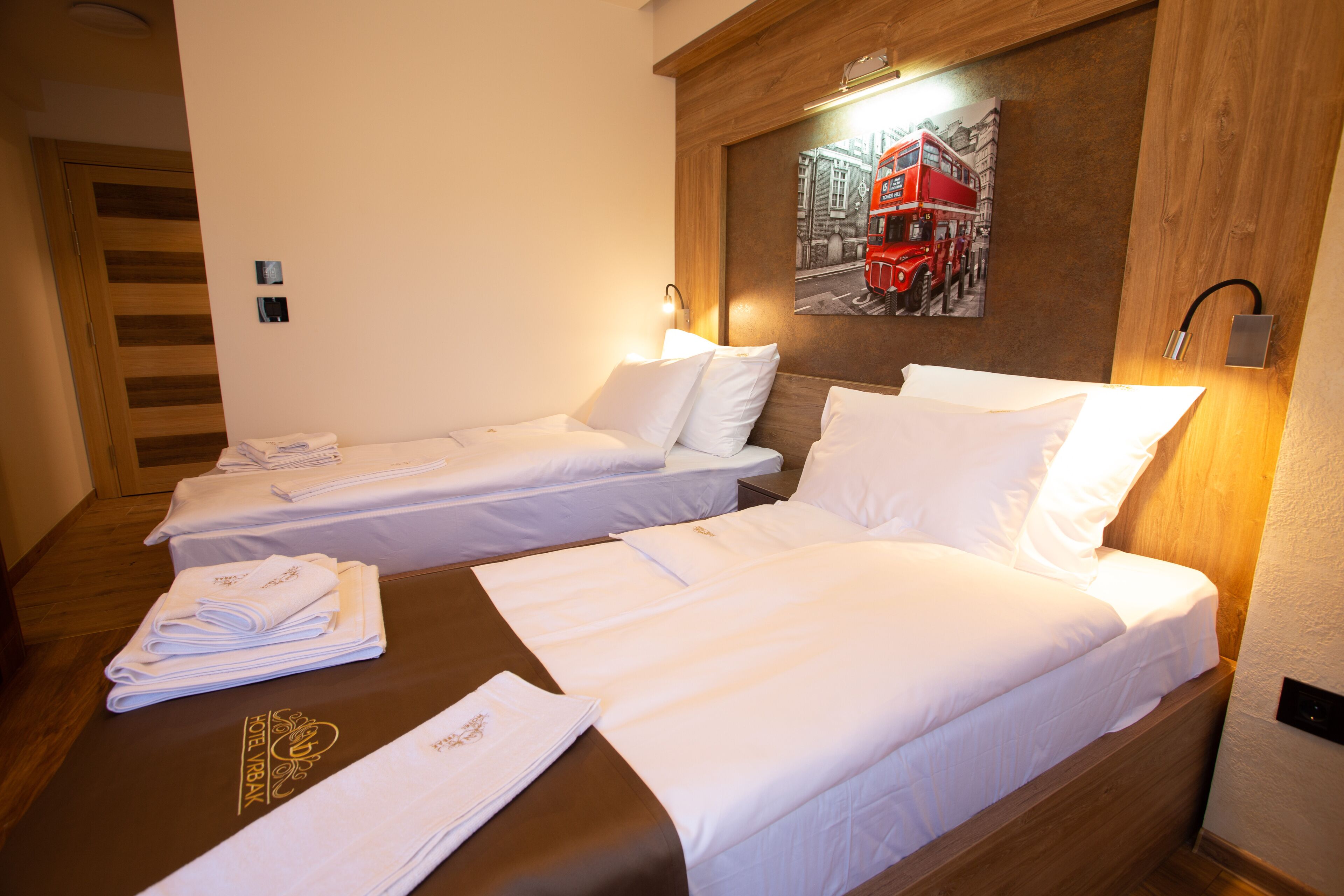 Quarto de hotel com duas camas individuais, cabeceira de madeira, obra de arte na parede retratando um ônibus de dois andares vermelho e toalhas dobradas no chão.
