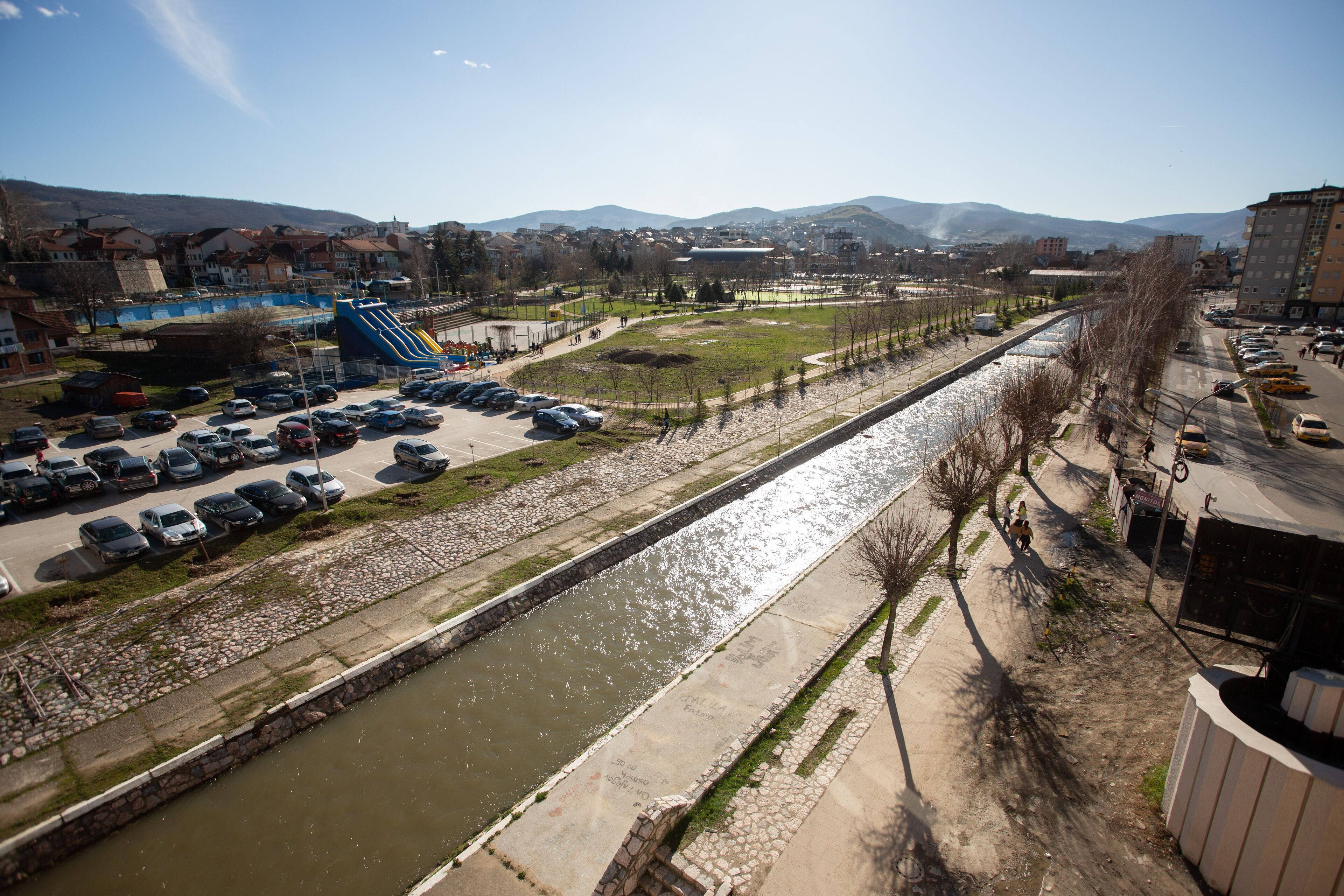Un río fluye junto a una carretera con coches aparcados y un parque infantil.