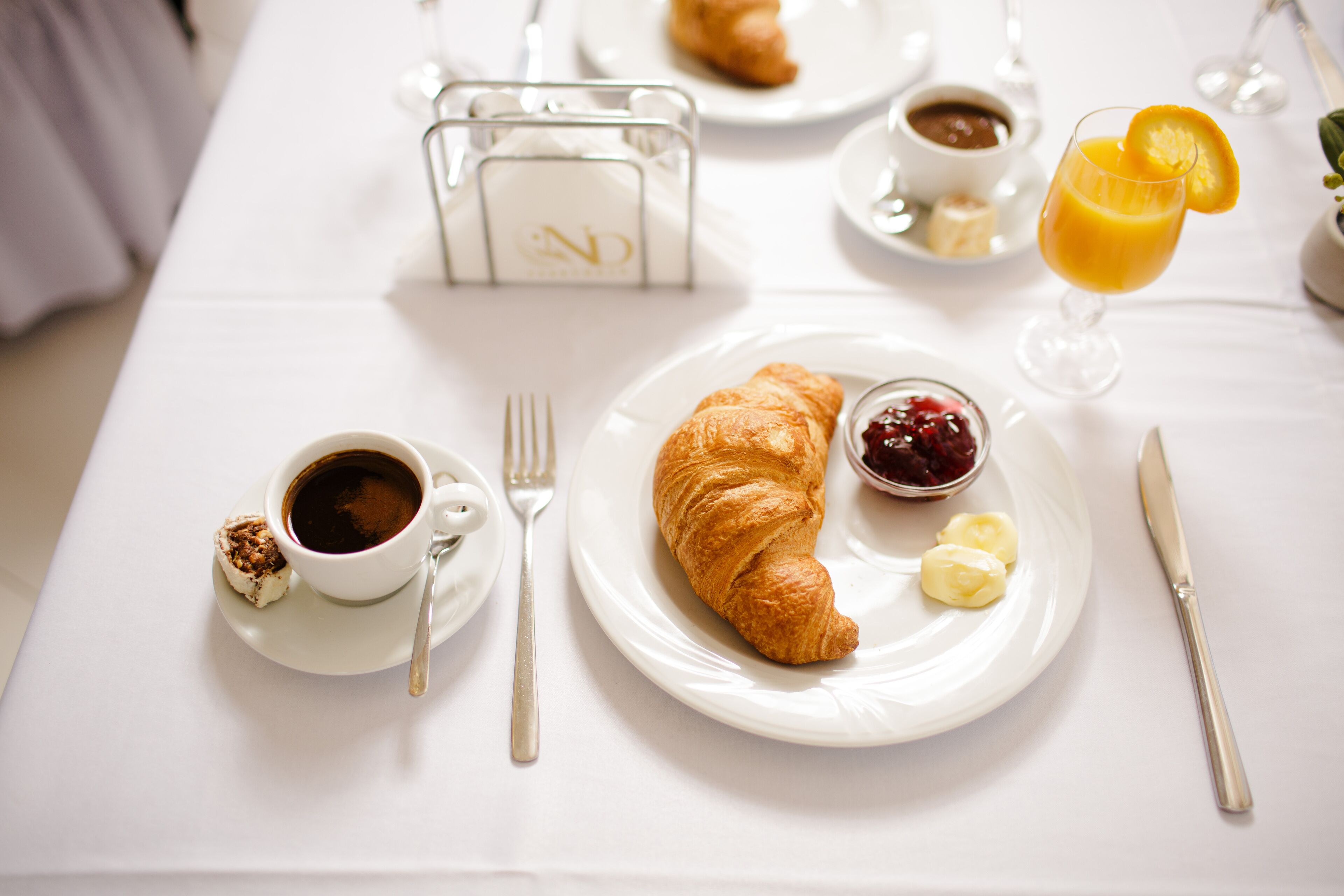 Desayuno con cruasanes, café, mantequilla, mermelada y jugo de naranja sobre una mesa blanca.