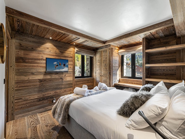 A wooden cabin bedroom with a large bed, a TV on the wall, and a window with a view of the outside.