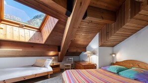 Een knus slaapkamer op zolder met een schuine plafond, houten balken, een groot raam en een kleurrijke gestreepte deken.