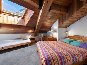 Ein gemütliches Schlafzimmer im Dachgeschoss mit schrägen Wänden, Holzbalken, einem großen Fenster und einer farbenfrohen, gestreiften Bettdecke.