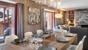 Comedor con una mesa de madera dispuesta para seis personas, rodeada de sillas. Se observa una pared de piedra y una chimenea al fondo.