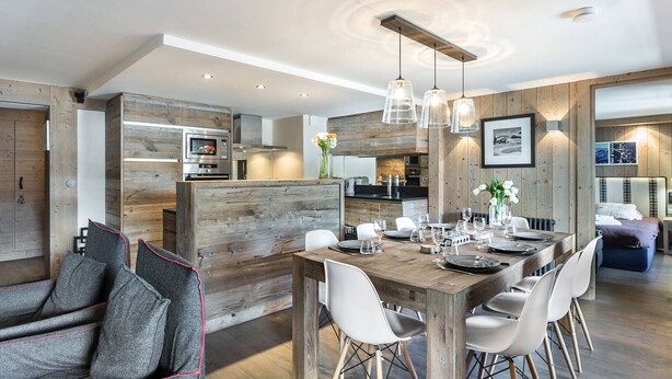 Una cocina moderna con detalles de madera, un comedor con mesa de madera y sillas blancas, y una zona de estar acogedora con sofá y cojines.