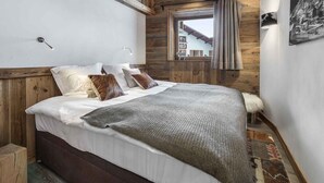 A bedroom with a wooden ceiling, a bed with white and brown bedding, a window with curtains, and a mounted picture on the wall. - Luxurious 4-Bedroom Apartment in Val d'Isère with Hammam, Fireplace, and Balconies (Val-d'Isère)