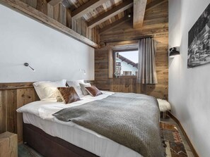 Un dormitorio con techo de madera, cama con ropa de cama blanca y marrón, ventana con cortinas e imagen colgada en la pared.