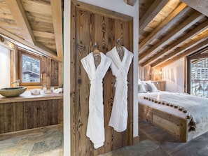 Interior de una cabaña de madera con una cama con estructura de madera, un tocador de madera con lavabo y dos albornoces blancos colgados en una puerta de madera.