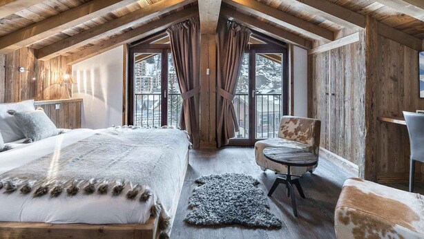 A cozy attic bedroom with a large bed, a chair, and a view of a snowy landscape through a window. - Luxurious 4-Bedroom Apartment in Val d'Isère with Hammam, Fireplace, and Balconies (Val-d'Isère)