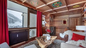 L’intérieur chaleureux d’un chalet avec un plafond en bois, une grande fenêtre ornée de rideaux rouges, une table basse en bois et un canapé agrémenté d’un plaid blanc et moelleux.
