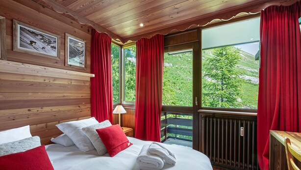 Une chambre dans un chalet en bois, avec un lit, des rideaux rouges et une vue sur la verdure environnante.