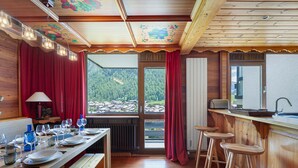 Un intérieur en bois comprenant une table à manger pour quatre personnes, un coin bar avec des tabourets et une vue sur un paysage boisé grâce à de grandes fenêtres.
