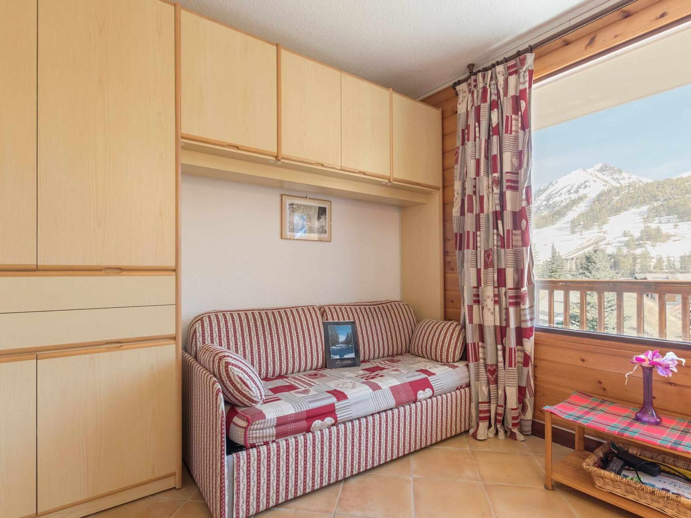 A cozy room with a striped sofa, wooden cabinets, a small table with a vase, and a window with a mountain view.