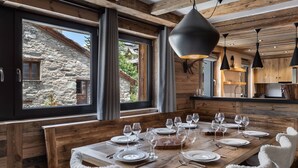 Una mesa de comedor de madera dispuesta con platos, vasos y cubiertos, con un edificio de piedra visible a través de amplios ventanales.