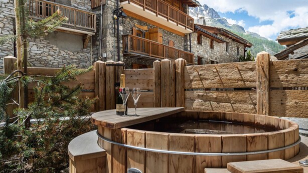Una bañera de hidromasaje de madera en un entorno rústico al aire libre, con edificios de piedra y montañas al fondo.