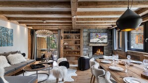 Un interior acogedor y rústico con chimenea de piedra, vigas de madera, un comedor con mesa dispuesta para seis personas y un televisor grande montado en la pared.
