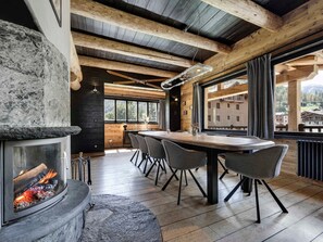 A modern interior with a stone fireplace, wooden beams, a dining area with chairs, and large windows. - Charming 200m² Chalet in Val d'Isère with Sauna and Nordic Bath (Val-d'Isère)