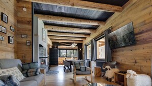 A log cabin interior with a fireplace, wooden floors, a dining area, and a large window.