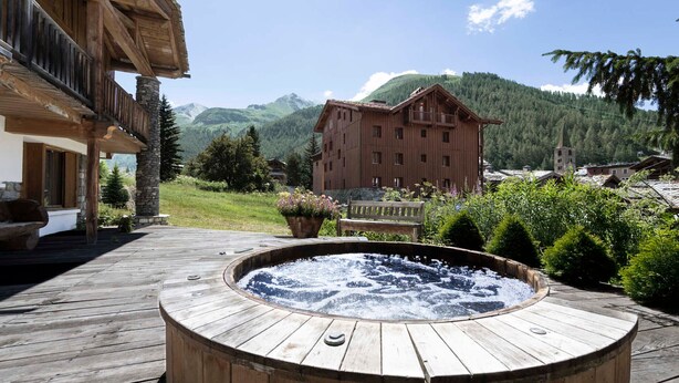 A wooden hot tub on a deck with a mountain view, a wooden building, and a stone pillar.