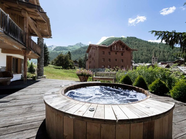 Un bain à remous en bois installé sur une terrasse avec vue sur les montagnes, un bâtiment en bois et un pilier en pierre.