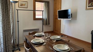 Un comedor con una mesa de madera dispuesta para cuatro personas, un televisor montado en la pared y un ventanal con cortinas.