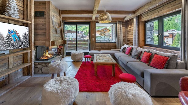 Ein gemütliches Wohnzimmer mit Kamin, Holzbalken, einem großen Fenster und einer Couch mit roten Kissen.