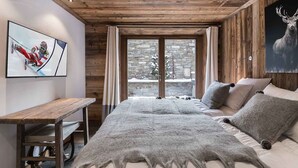 Une chambre confortable avec un plafond en bois, une grande fenêtre donnant sur le paysage enneigé, un lit recouvert d’un plaid en fourrure et un bureau avec une chaise.