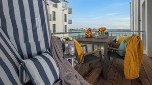 Un balcone con un divano a righe, un tavolo con sedie, un vaso con fiori e vista sul mare.