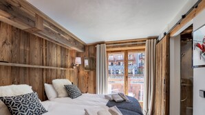 Habitación de hotel con paredes de paneles de madera, cama con almohadas blancas y estampadas, una mesita con lámpara y vistas al balcón exterior.