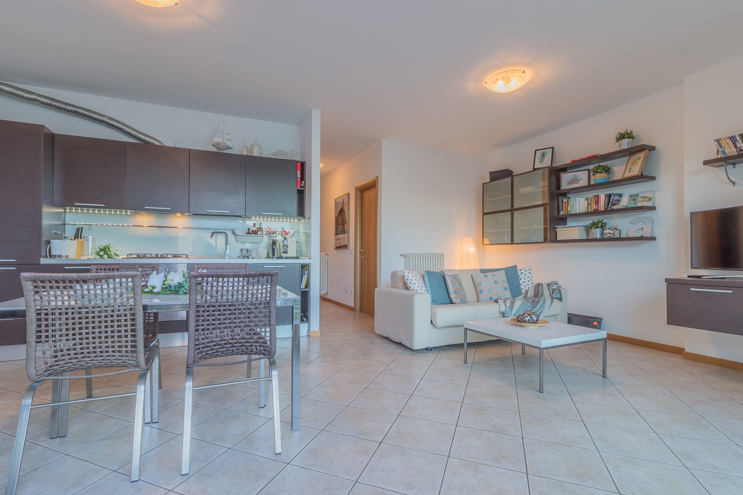 Una sala moderna con un sofá blanco, dos sillas de mimbre, una mesa de centro y una cocina con mesa de comedor.