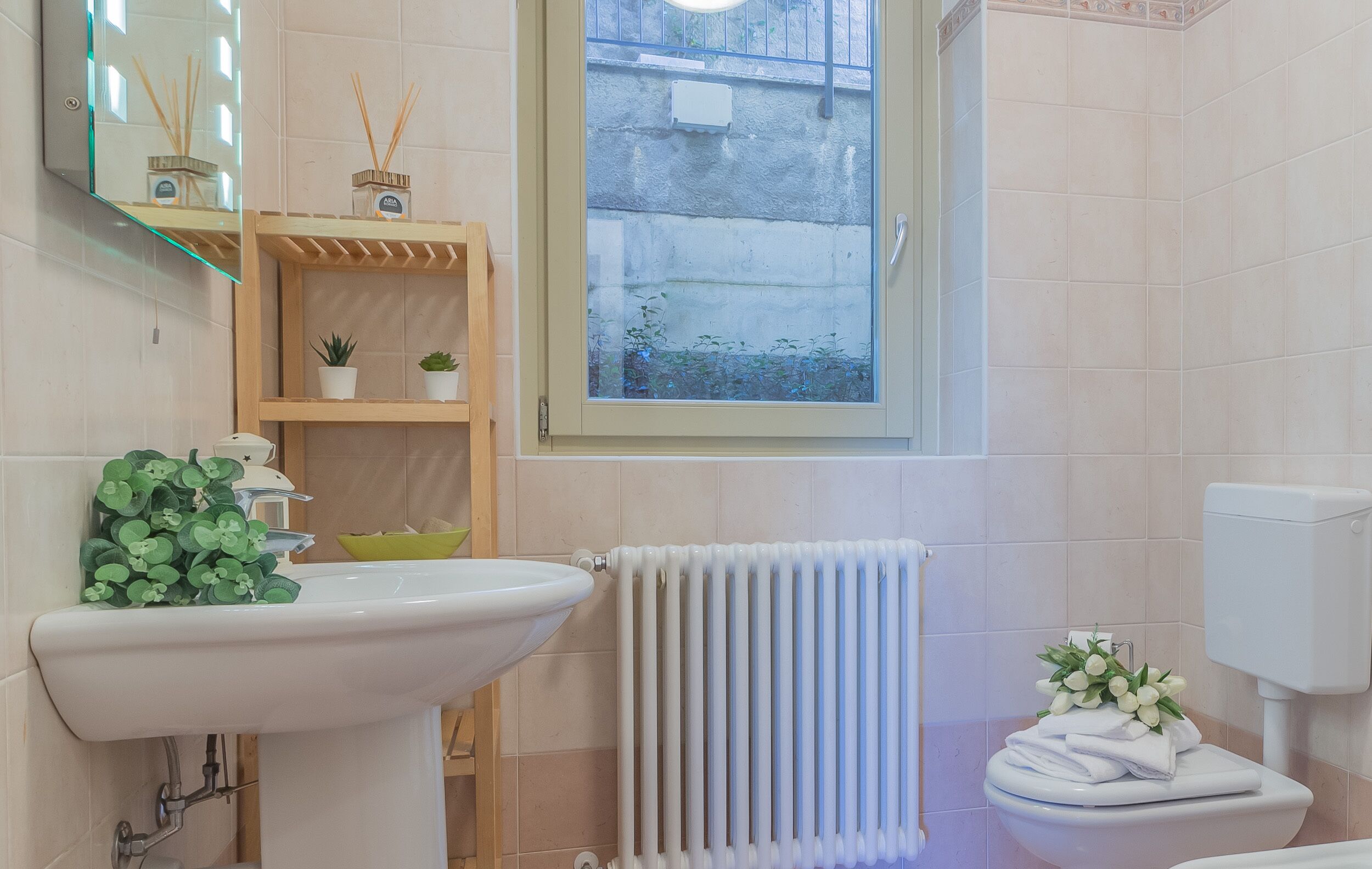 A bathroom with a white sink, a window with a view of a building, a radiator, and a toilet with a floral decoration.