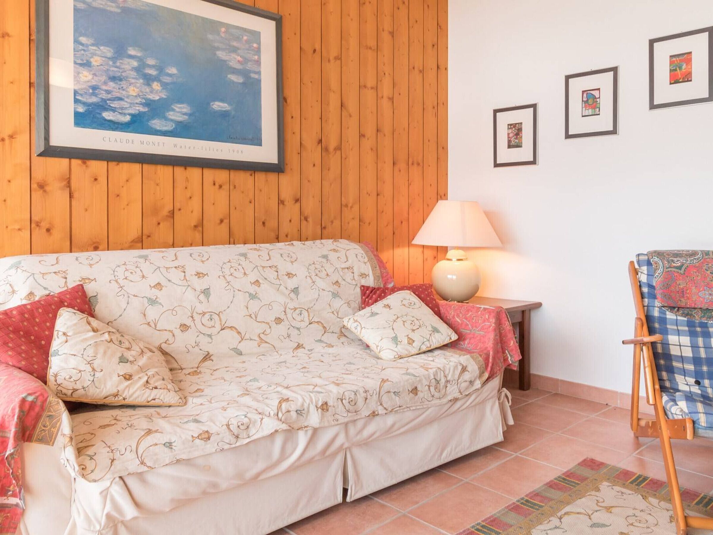 Een knus interieur met een houten wand, een schilderij van waterlelies in een lijst, een bank met bloemenkussens, een bijzettafeltje met een lamp en een stoel met een gestreept kussen.