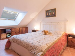 Quarto com teto inclinado, claraboia, cama com colcha estampada, mesa de cabeceira de madeira, abajur e cômoda de madeira pequena.