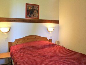 Quarto com cabeceira de madeira, colcha vermelha, duas luzes na parede e um quadro emoldurado sobre a cama.