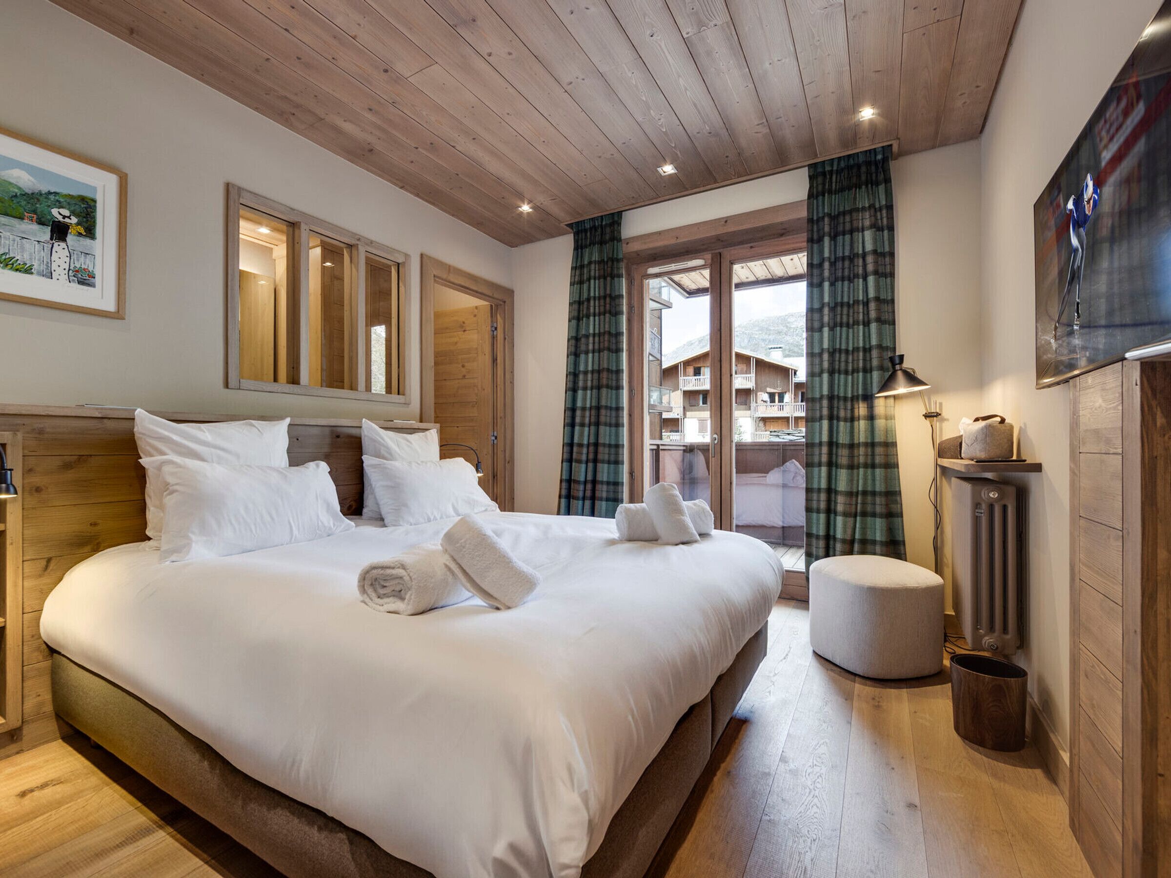 Un dormitorio con una cama grande, cabecera de madera, una mesita de noche, una lámpara y vistas a un edificio a través de una ventana con cortinas.