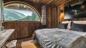 A cozy wooden cabin bedroom with a large bed, a dining table, and a scenic mountain view through a large arched window. - Luxurious Chalet with Panoramic Views, Jacuzzi, Hammam, Near Slopes, Sleeps 10 (Val-d'Isère)