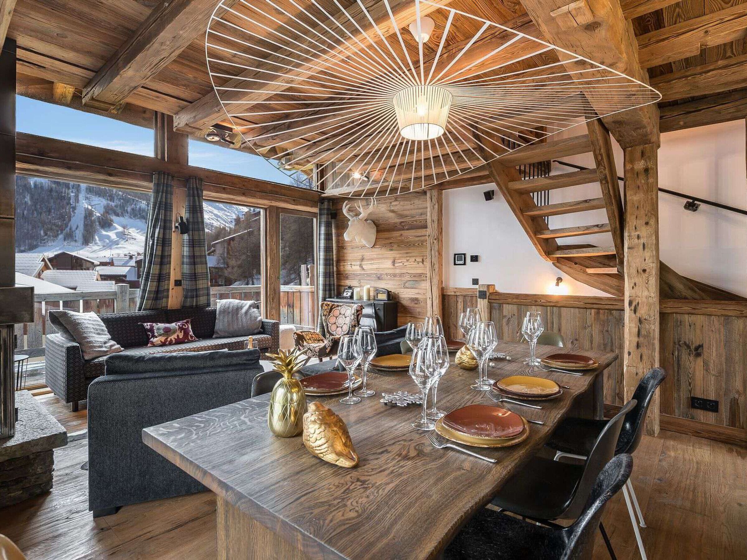 Un interior de madera con una mesa de comedor para seis comensales, un sofá y un ventanal con vistas a montañas nevadas.
