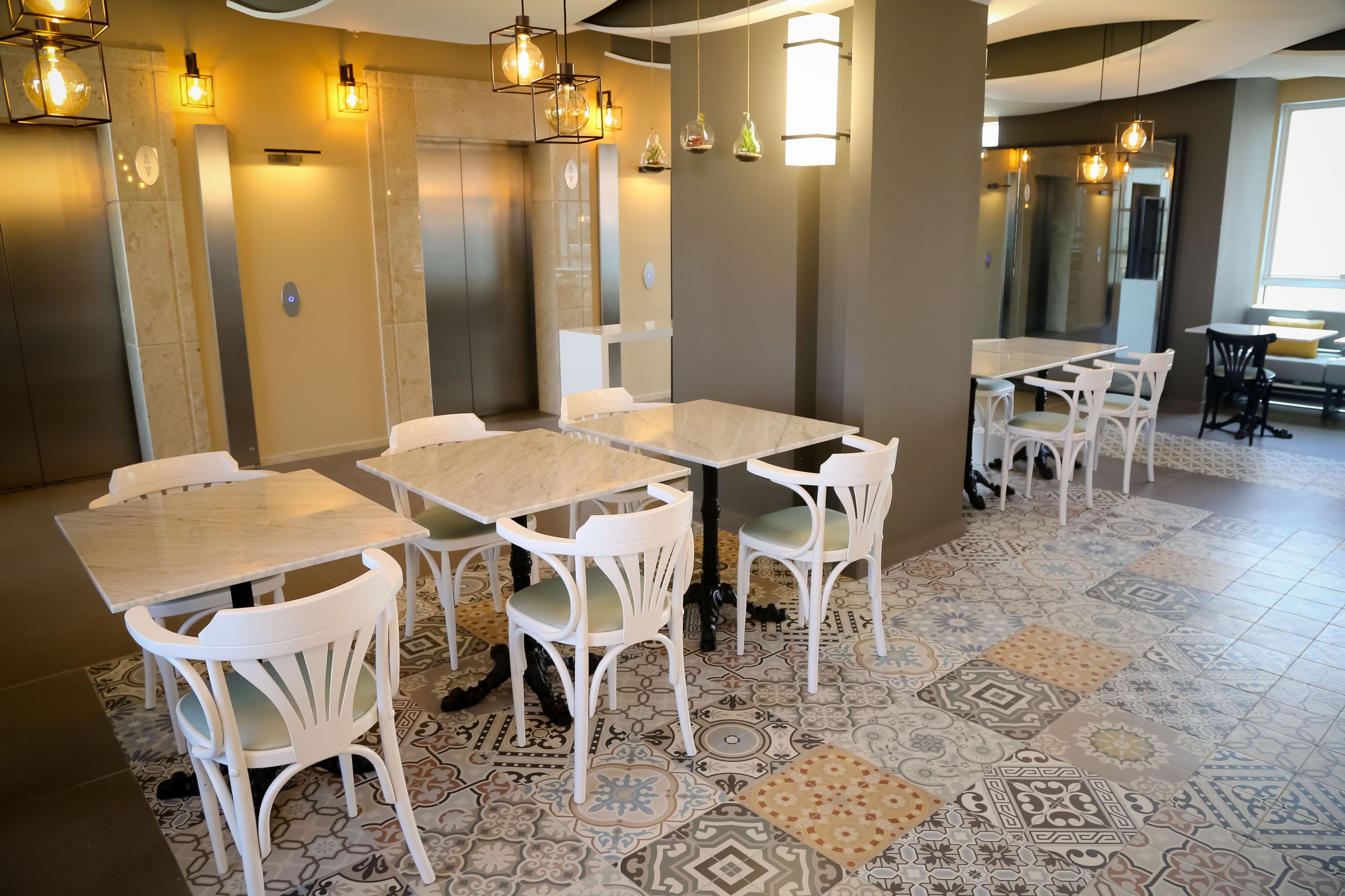 Een moderne eetruimte met witte stoelen en tafels, vloertegels met een patroon en hanglampen.