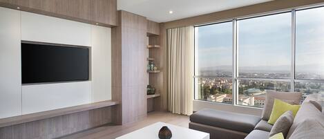 Ein modernes Wohnzimmer mit einem an der Wand befestigten Flachbildfernseher, einem großen Fenster mit Blick auf die Stadt, einem grauen Sofa mit gelben Kissen und einem weißen Couchtisch.