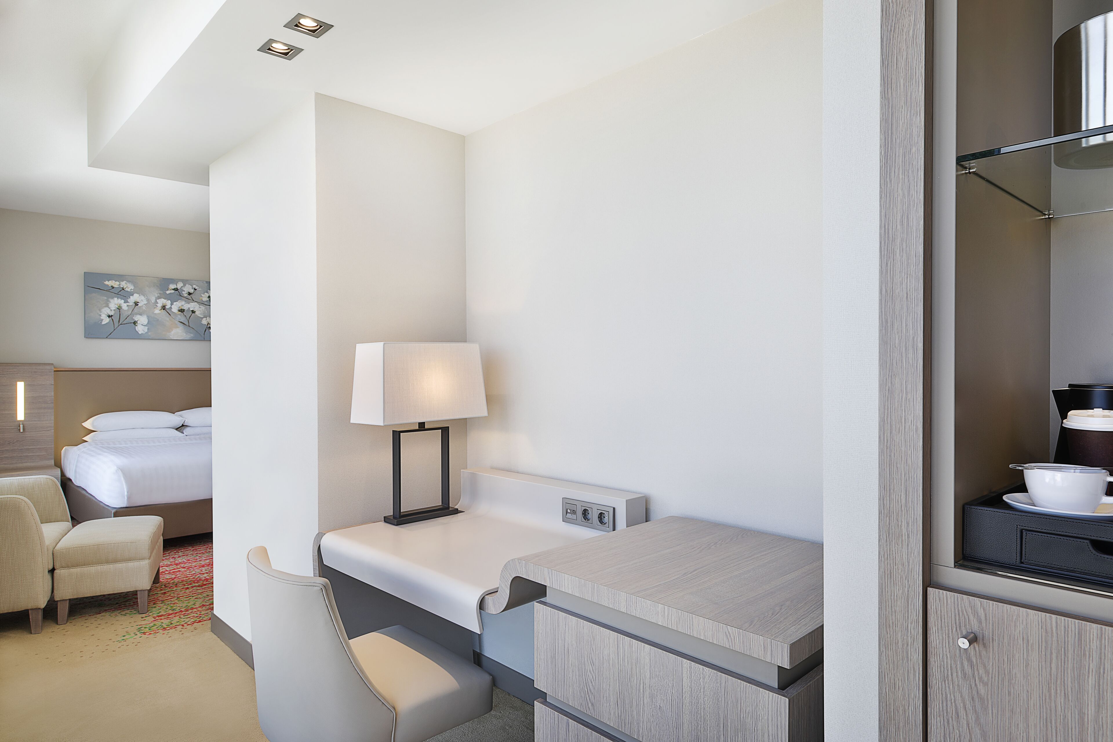 A hotel room with a bed, a desk with a lamp, a chair, a small table with a cup and saucer, and a painting on the wall.